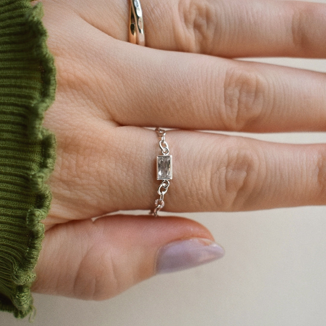 A silver ring with a rectangular clear gemstone set in a chain-link band, worn on a hand with lavender nail polish and an olive green sleeve visible.