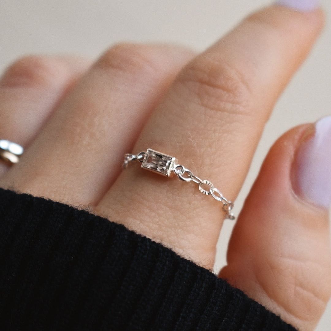 A close-up of a hand wearing a delicate silver chain ring with a small rectangular clear gemstone, alongside another silver band ring on another finger.