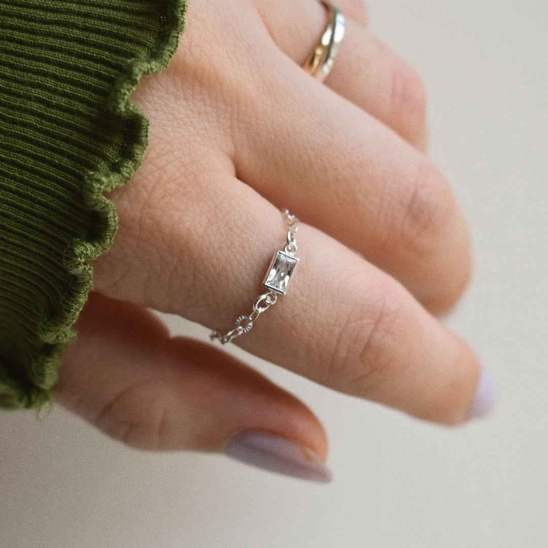 A close-up of a hand wearing a delicate silver chain ring with small clear stones, alongside an olive green ribbed sleeve, against a soft off-white background.