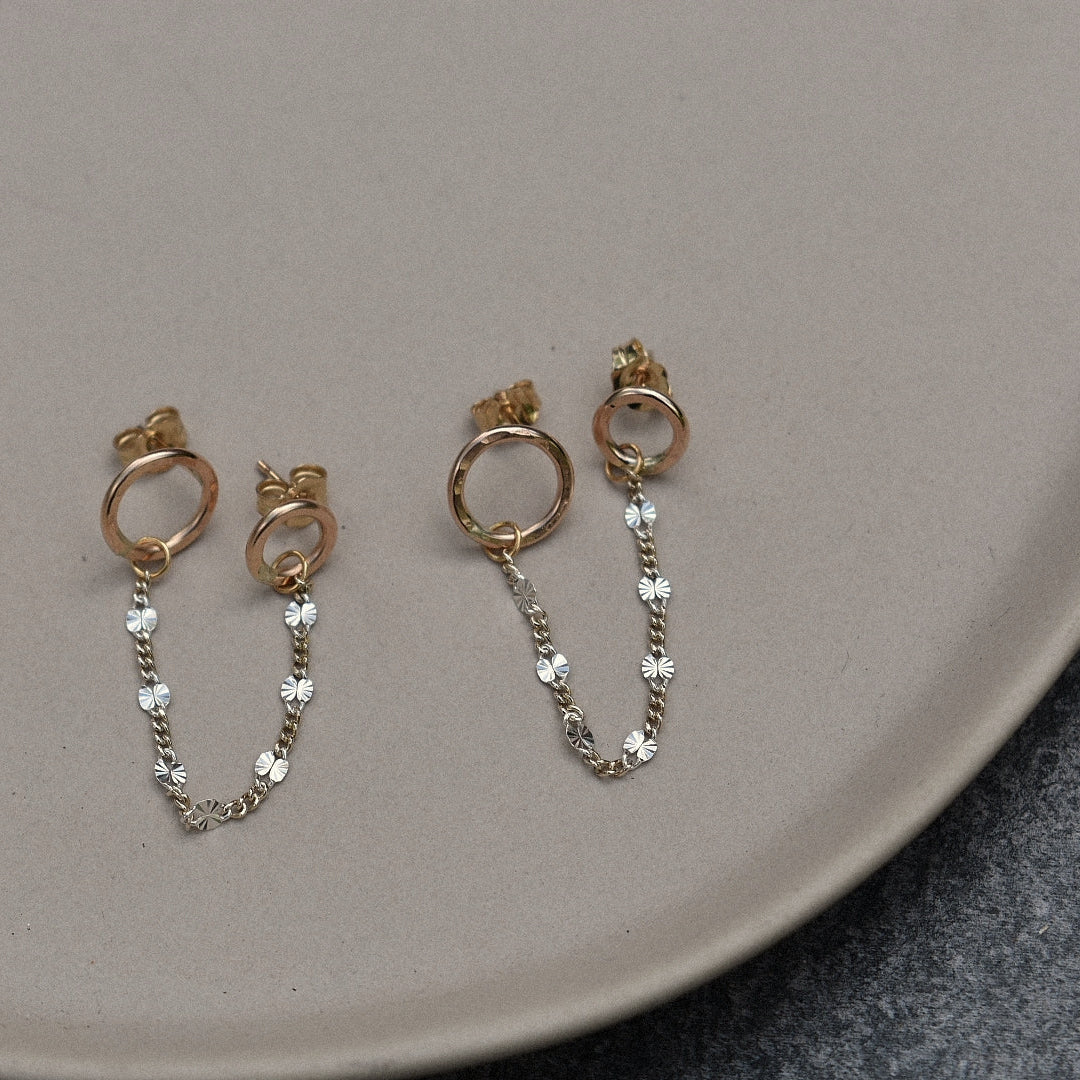 A pair of gold-toned dangle earrings with circular hoops and short chains of clear rhinestones, displayed on a beige ceramic tray.